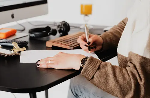 Femme avec du vernis noir en train d'écrire sur une feuille devant son ordinateur à son bureau